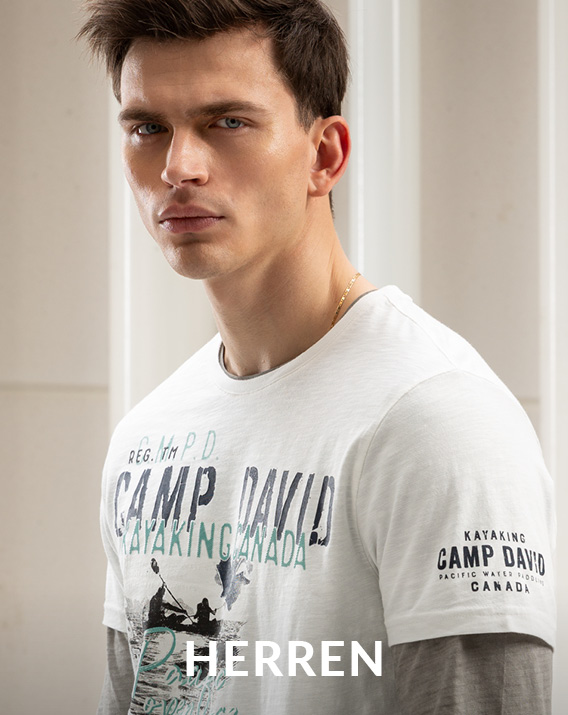 Männermodel mit cremefarbenen Hoodie und kurzes Skaterjeans lehnt an einer Reling auf einem Motorboot und blickt nach rechts.