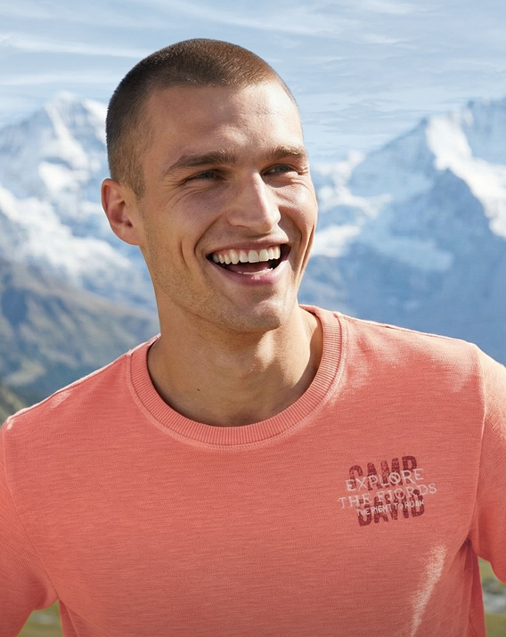 Model steht in einer Berglandschaft und trägt eine weiße Jacke