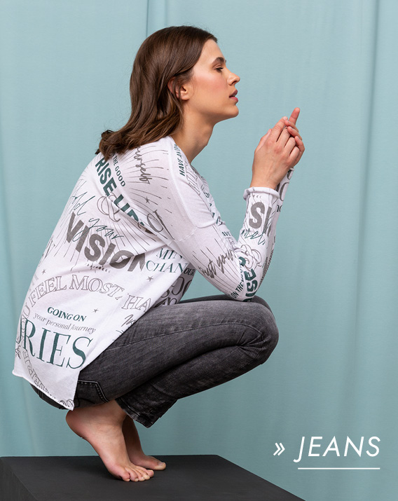 Model trägt schwarze Jeans und ein weißes Oberteil und hockt auf einem Hocker